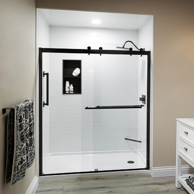 Bath Concepts Prime Bath shower with white subway-texture acrylic walls, a matte black Denali barn-style sliding glass door, niche and fold-down seat