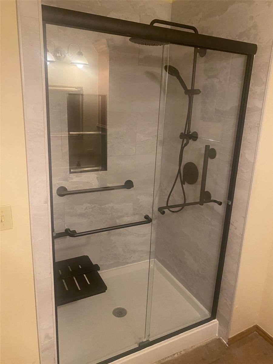 Customer's shower as photographed: gray stone-look wall panels, black-framed sliding glass door, grab bars, fold-down seat, and a black rain shower column